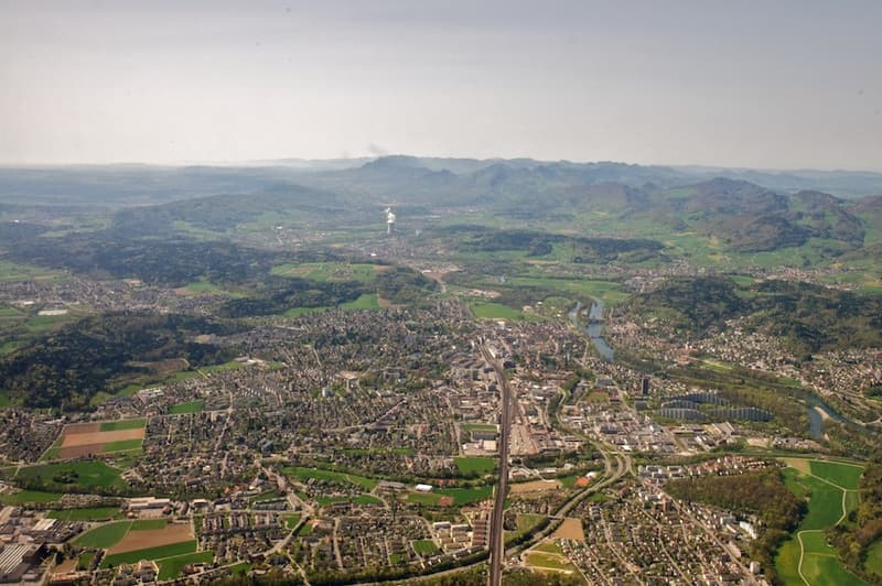 An aerial view of a cityscape surrounded by green fields and hills, with a winding river and distant mountains.