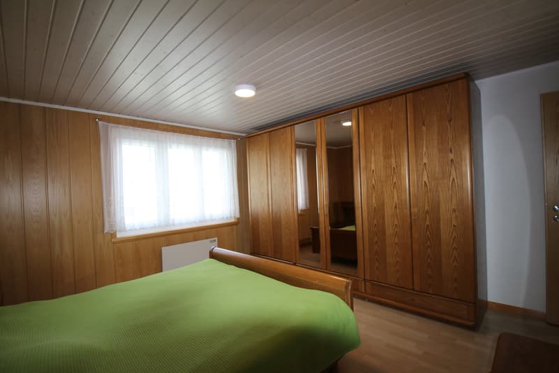large wooden wardrobe, double bed, white curtains on window, wooden floor, beige wall, radiator