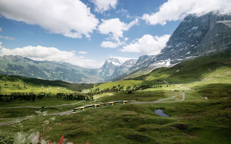 A train is traveling through a mountainous valley with lush green fields and a clear blue sky.