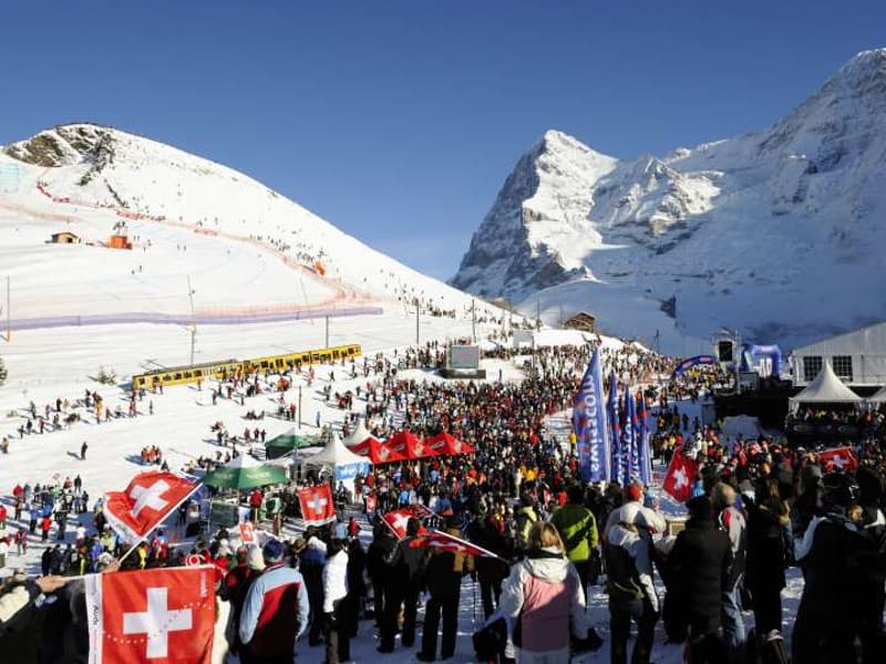 Crowd of people gathered for a skiing event, flags, tents, mountains in the background