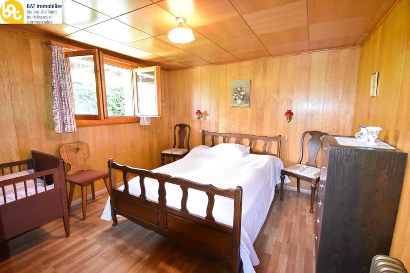 Small bedroom with wooden walls, double bed, wooden chair, small window with curtains, wooden chest of drawers