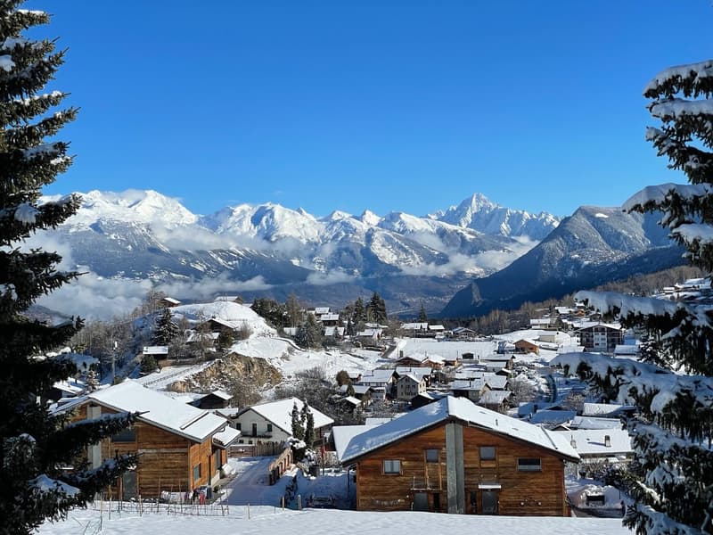 Snowy village with wooden houses, mountains in the background, clear blue sky, pine trees