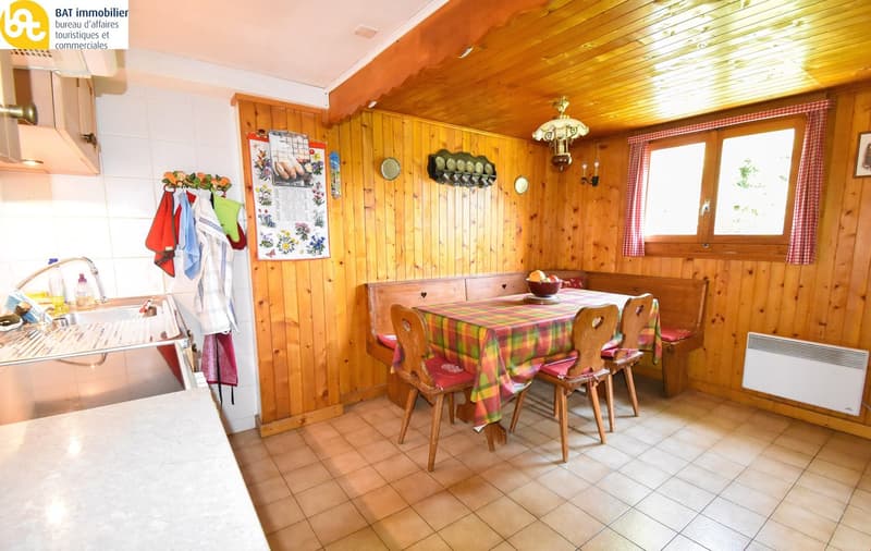 A wooden-walled kitchen with a white countertop, tile floor, a stainless steel sink, and a hanging oven above the sink. A dining table with colorful cushions and wooden chairs. A wooden bench seat on the right side, a wooden cabinet on the left, a framed