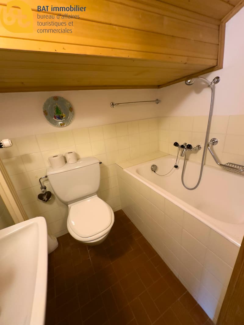 Bathroom with toilet, bathtub, and shower. Tiled walls and floor. Wooden ceiling. Shelves for storage.