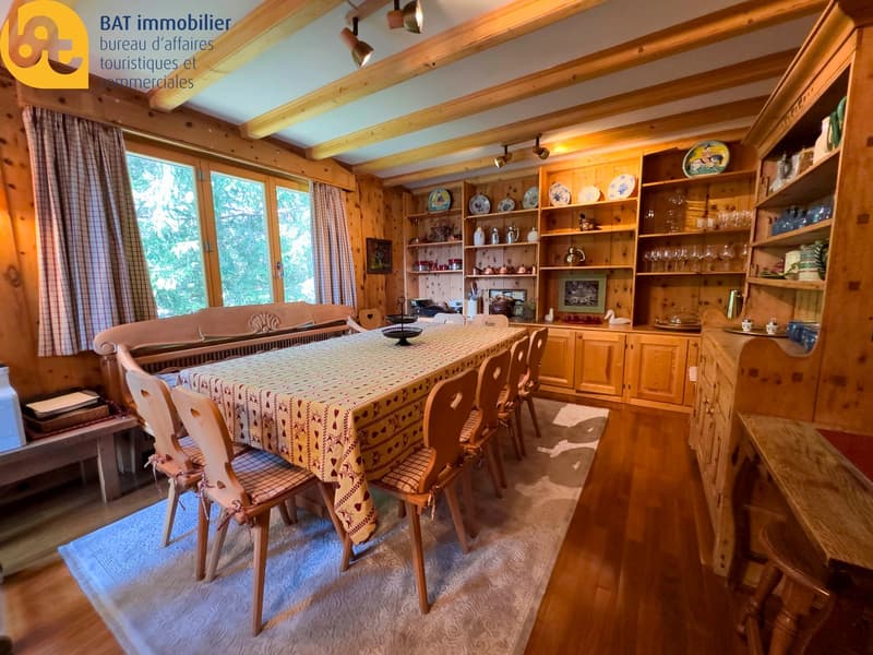 Spacious dining room with wooden beams, large windows, wooden furniture, shelves displaying decorative items, and a patterned tablecloth on the dining table.