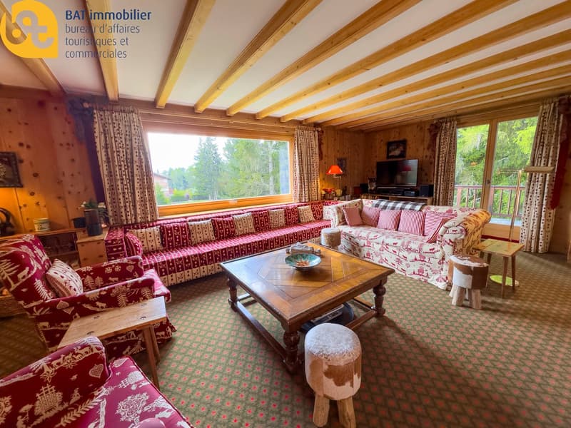 Cozy living room with wooden beamed ceiling, large windows overlooking the outdoors, patterned upholstered sofas and chairs, wooden coffee table, and decorative accents.