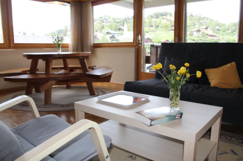 Spacious living room with large windows offering a scenic view of the surrounding area. The room features a wooden dining table with benches, a black sofa, a white coffee table, and a vase of yellow flowers adding a touch of color. The overall decor has a