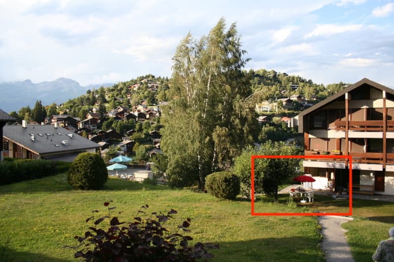 Scenic mountain village with wooden chalets, surrounded by lush greenery and trees. The image shows a building with a balcony and a parking area in the foreground.