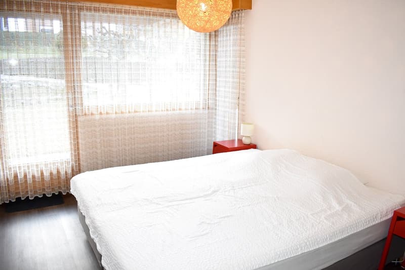 Bedroom with large window, white bedding, red side table, and a decorative light fixture