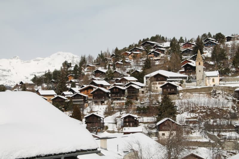 A picturesque snow-covered village nestled in the mountains, with traditional Swiss-style chalets and a church steeple visible. The buildings are surrounded by pine trees and the landscape is blanketed in snow.