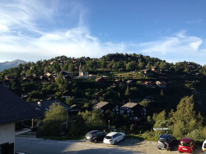 The image shows a scenic mountain village with a church steeple visible. The village is surrounded by lush green forests and hills, with a clear blue sky overhead. There is a parking area visible in the foreground with several cars parked.