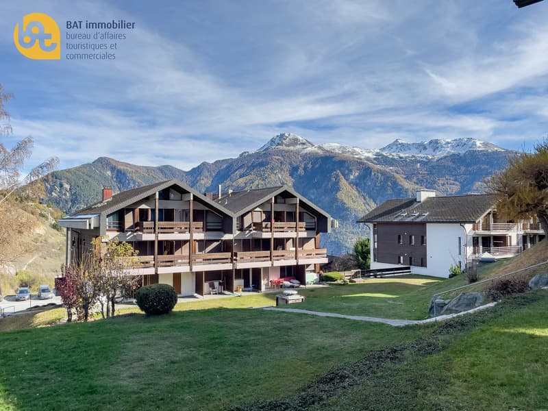 Multi-story apartment building with wooden exterior, balconies, surrounded by a grassy lawn, with a scenic mountain view in the background