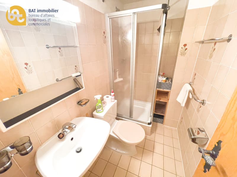 Tiled bathroom with a shower enclosure, toilet, sink, and storage shelves. The bathroom appears to be equipped with a private washing machine.