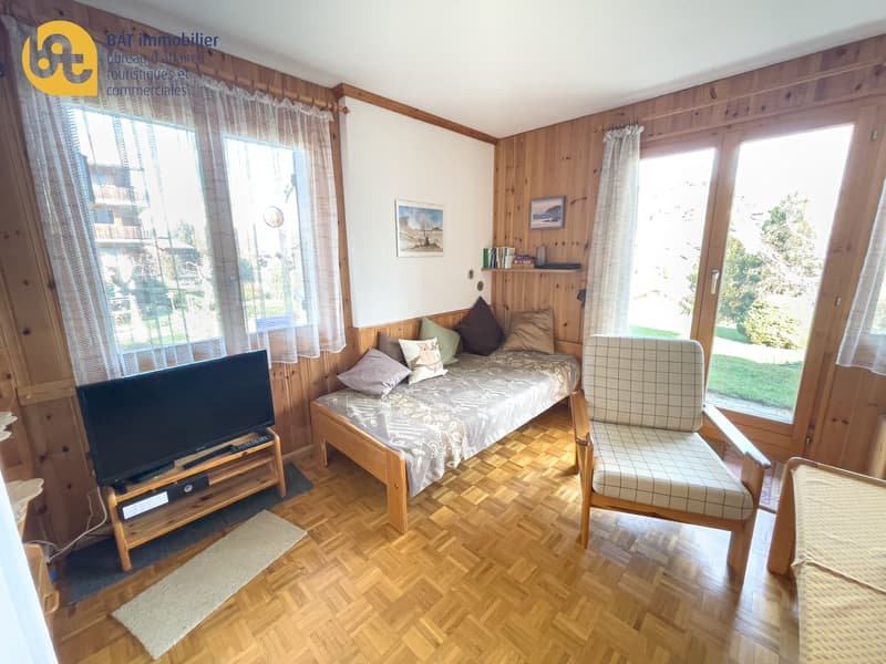 Cozy bedroom with wooden walls and floors, large windows, a double bed, a TV stand, and a patterned armchair.