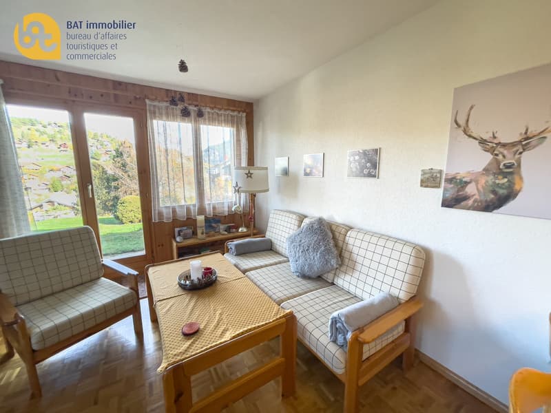 Cozy living room with large windows overlooking a garden, wooden furniture including a sofa and coffee table, wall decor with a deer painting, and a balcony or terrace visible through the windows.