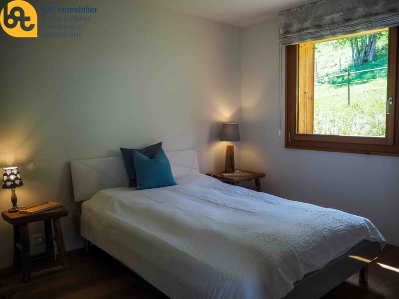 Cozy bedroom with large window overlooking a lush, green outdoor area. The room features a comfortable bed with white bedding and blue accent pillows, a wooden side table with a lamp, and a wooden window frame.
