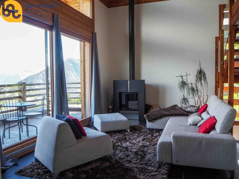 Spacious living room with large windows, wooden ceiling, gray sofas, red accent pillows, fireplace, and access to a balcony or terrace with a view of the mountains