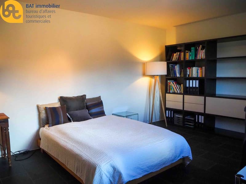 Bedroom with a double bed, dark colored bedding, a bookshelf with books and binders, and a lamp on a side table.