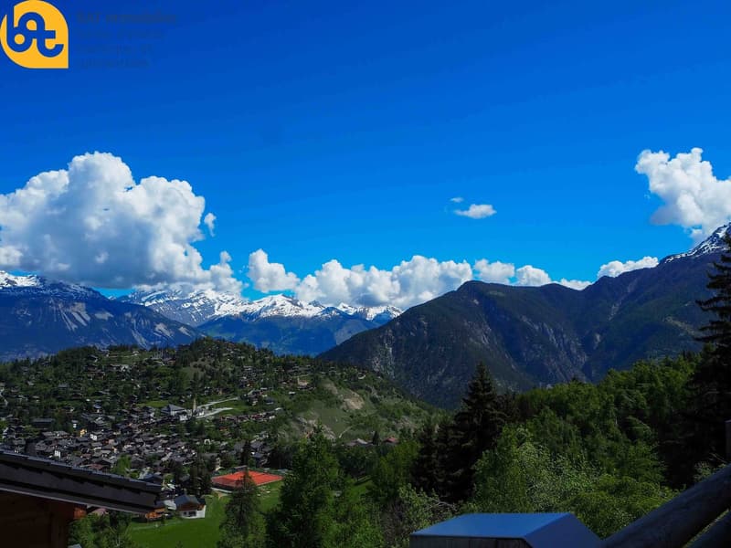 The image shows a scenic mountain landscape with a small town nestled in the valley below. The mountains are covered in snow and there are fluffy white clouds in the bright blue sky. The town is surrounded by lush green forests and the overall scene is pi