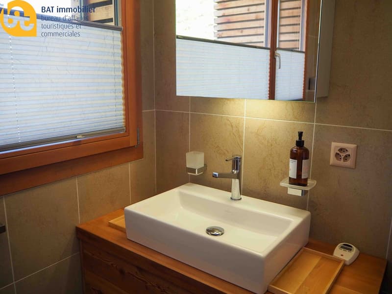 Bathroom with a white ceramic sink, wooden vanity, tiled walls, and a window with blinds.