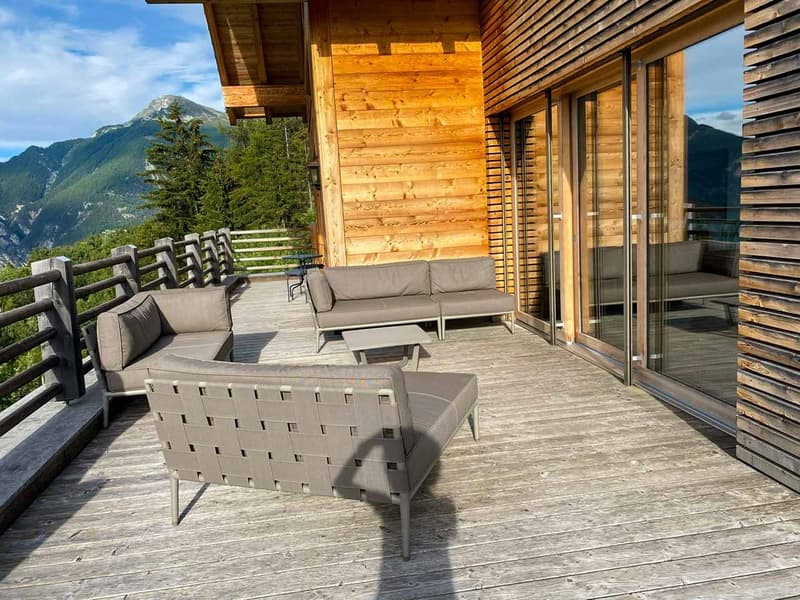 Wooden deck with outdoor furniture, including a sofa and coffee table, overlooking a scenic mountain landscape with trees in the foreground