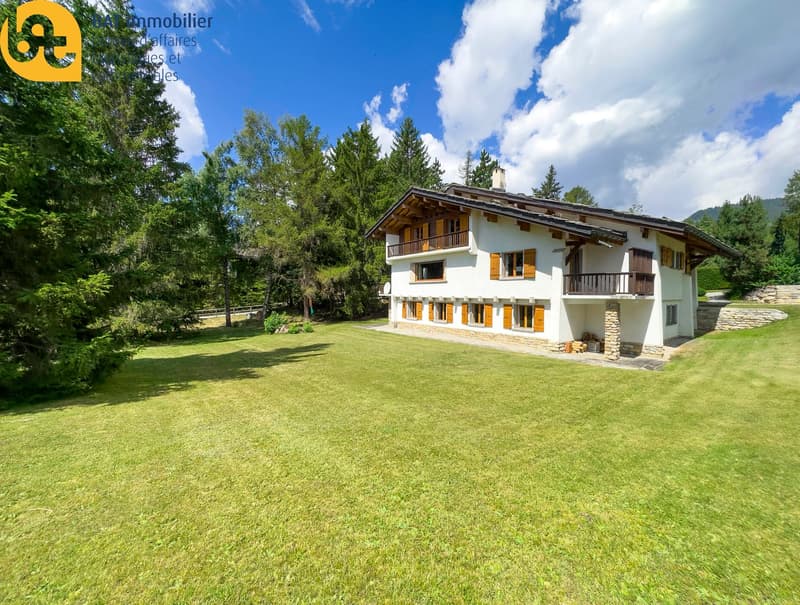 2-story chalet-style house with white exterior, wooden balconies, surrounded by lush green trees and lawn, with a paved parking area