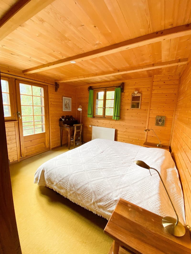 A bedroom with wooden walls and flooring. The bed has a white quilt. There are two lamps and a wooden desk with a chair. A radiator and picture frames are on the wall. The room has a glass window with green curtains.
