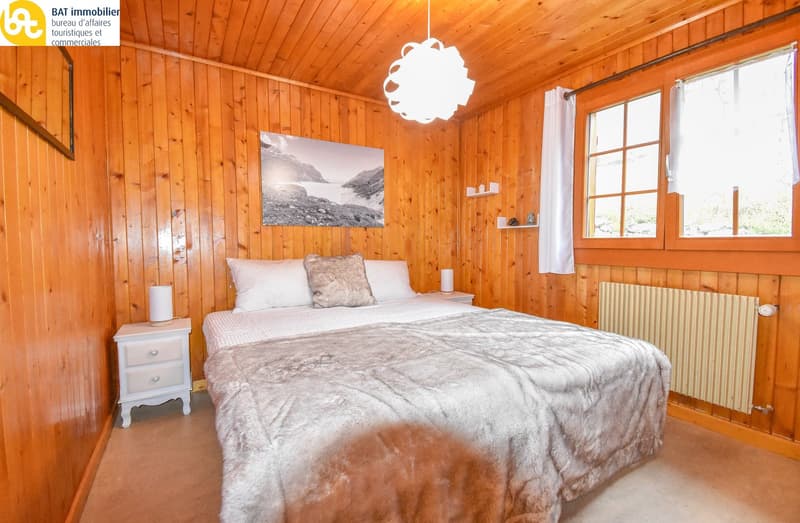 Cozy wooden bedroom with a large bed, fur blanket, and a landscape painting on the wall. The room has wooden paneled walls and a pendant light fixture.