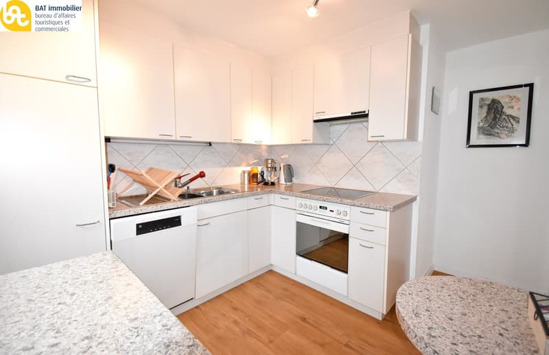 Modern, L-shaped kitchen with white cabinets, granite countertops, and tiled backsplash. Appliances include a stove, oven, and dishwasher. The kitchen has a wooden floor and appears to be part of a larger living space.