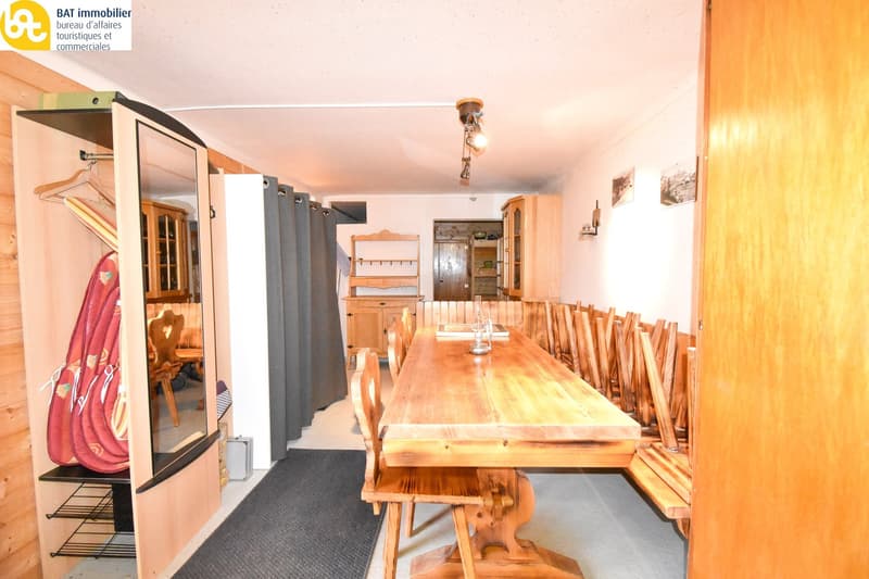 The image shows a dining room with a large wooden dining table and benches. There is a shelving unit on the wall and a closet or wardrobe visible. The room has a rustic, wooden interior design.
