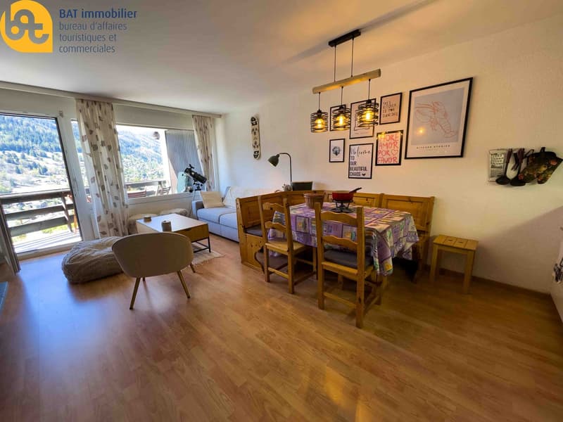 Spacious living room with large windows offering a scenic view, wooden dining table and chairs, sofa, and decorative wall art and lighting fixtures.