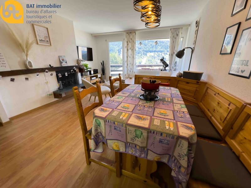 Open-plan living room and dining area with large windows offering a scenic view, wooden furniture including a dining table with a patterned tablecloth, and a cozy seating area with a TV and decorative elements.