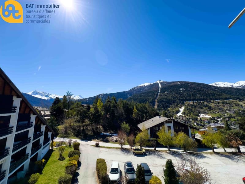 The image shows a scenic mountain landscape with snow-capped peaks, a ski resort with buildings and a parking area, and a clear blue sky with the sun shining brightly.