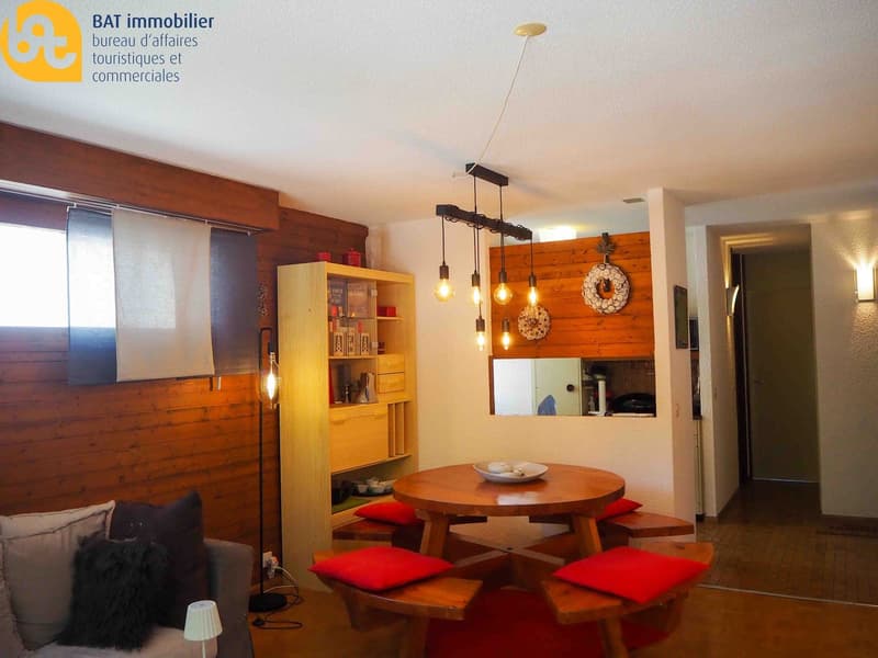 Cozy living room with wooden walls, round wooden dining table with red cushions, shelving unit, and hanging light fixtures