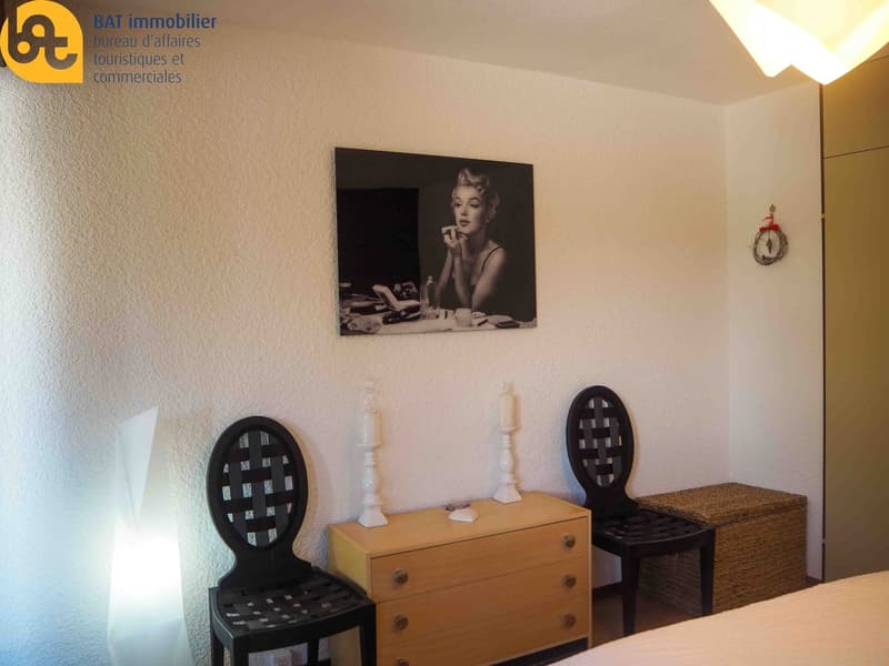 The image shows a room with white walls, a large black and white portrait of a woman on the wall, two black wicker chairs, a wooden dresser, and a wicker basket. The room has a cozy, vintage-inspired decor.