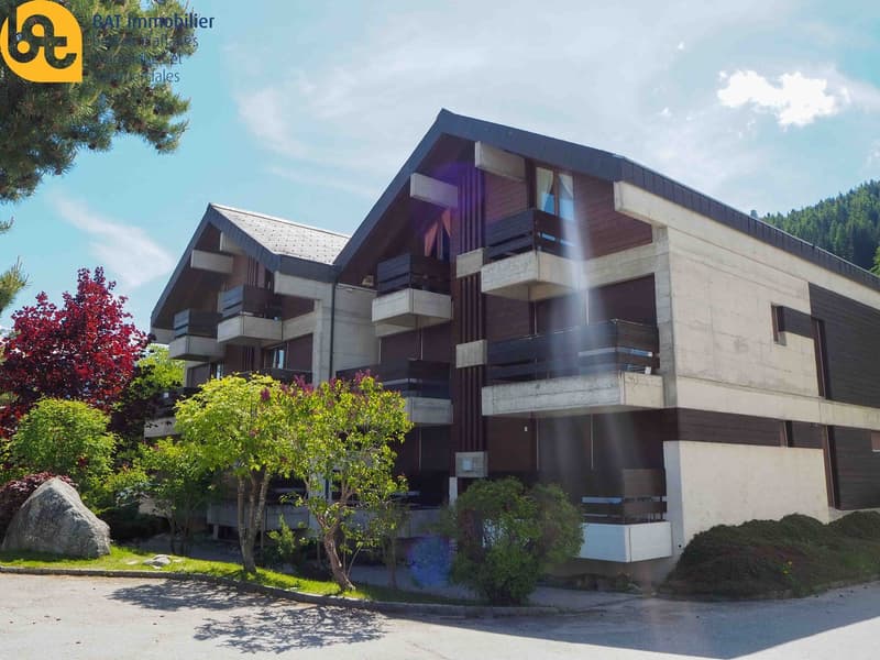Modern multi-story building, beige and brown facades, balconies, glass windows, trees, bushes, green lawn, mountain landscape