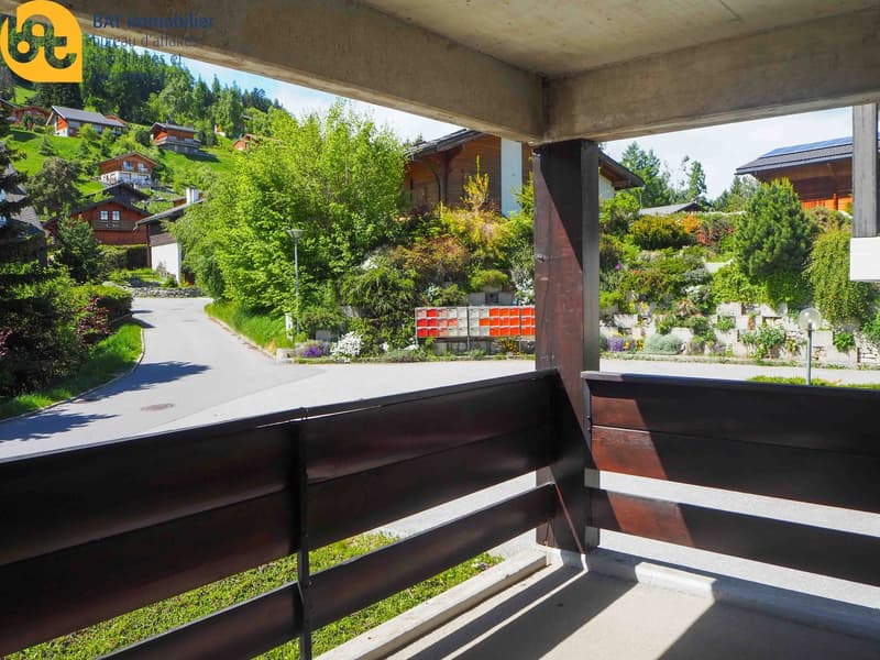 The image shows a balcony or terrace overlooking a scenic, forested area with wooden chalets or cabins in the background. The balcony has a wooden railing and appears to be part of a larger residential or vacation property.