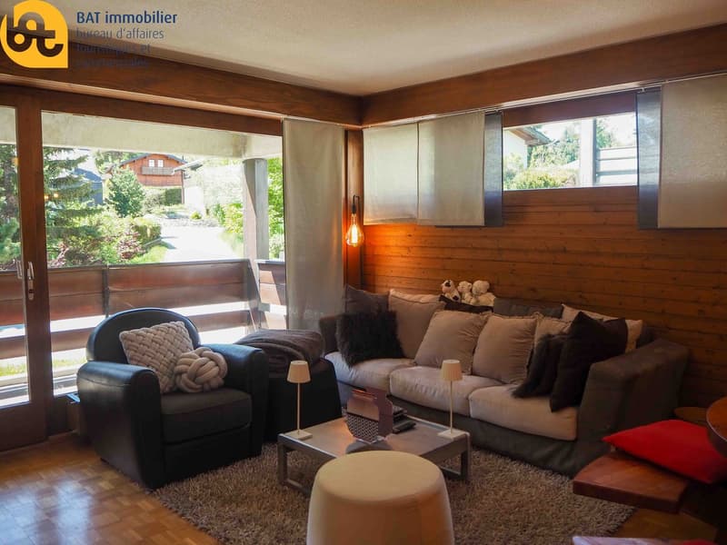 Cozy living room with large windows overlooking a garden, wooden paneled walls, comfortable sofa and armchair, coffee table, and decorative lighting fixtures.