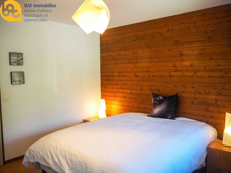 Cozy bedroom with wooden wall paneling, a large bed, and a black cat sitting on the bed. The room has a warm, inviting atmosphere with a fireplace visible.