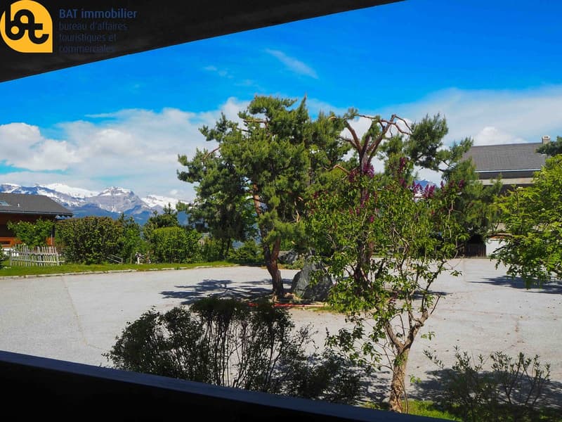 The image shows a scenic outdoor landscape with a large tree in the foreground, surrounded by lush greenery and shrubs. In the background, there are snow-capped mountains visible under a blue sky with some clouds. The scene appears to be in a mountainous,