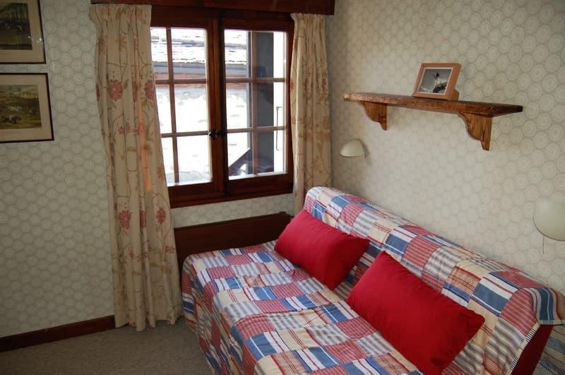 Cozy bedroom with patterned curtains, wooden window frames, a wooden shelf, and a plaid patterned bedspread with red accent pillows.