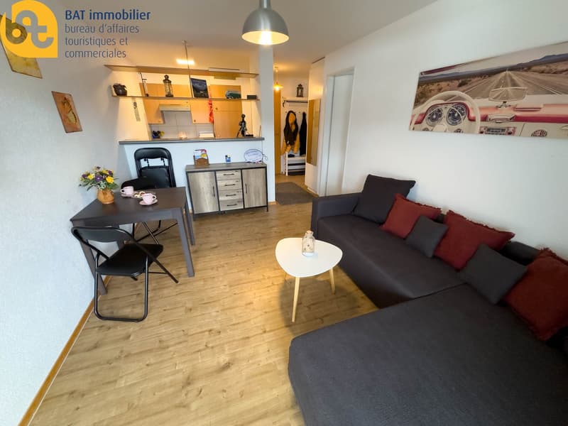 Open-plan studio apartment with hardwood floors, a dining table, a sofa, shelving units, and a kitchenette with a dishwasher. The room is decorated with various artwork and decor items.