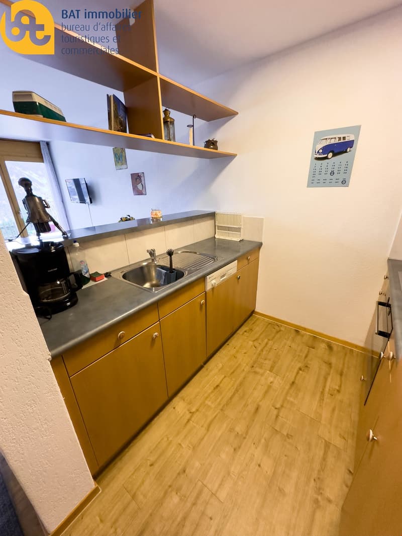Compact kitchen with yellow cabinets, stainless steel sink, and a dishwasher. Shelves above the cabinets display decorative items. The floor is made of wood-like laminate material.