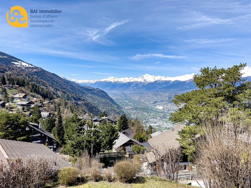 The image shows a scenic mountain landscape with snow-capped peaks in the distance. The foreground features a village nestled among the trees, with houses and buildings visible. The sky is clear and blue, with some wispy clouds. The overall scene depicts