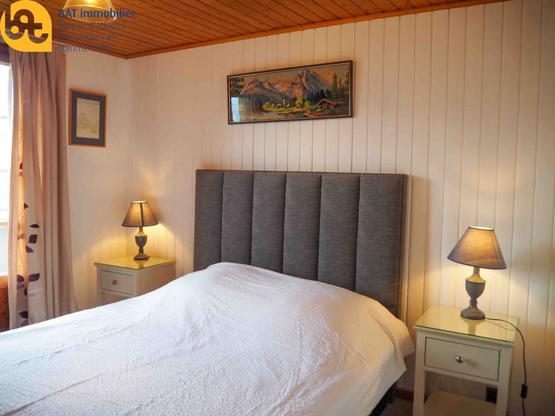 Cozy bedroom with wooden paneled walls, a large framed landscape painting, a grey upholstered headboard, a bedside table with a lamp, and curtains on the window.
