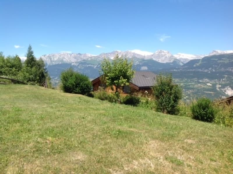 The image shows a scenic mountain landscape with a grassy field in the foreground and several wooden cabins or chalets nestled among the trees. In the background, there are snow-capped mountains visible under a clear blue sky.