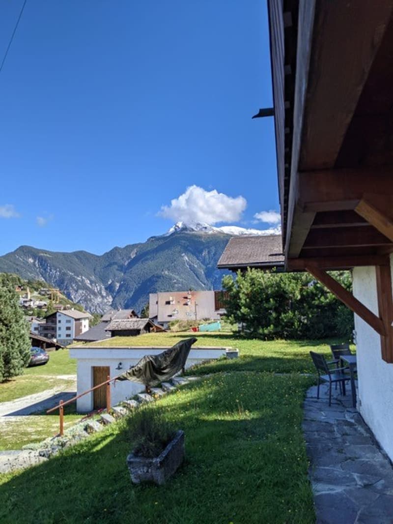The image shows a scenic mountain landscape with a village nestled in the valley below. The foreground features a grassy area with a wooden structure, likely a balcony or terrace, offering a panoramic view of the mountains and the village. The sky is a vi