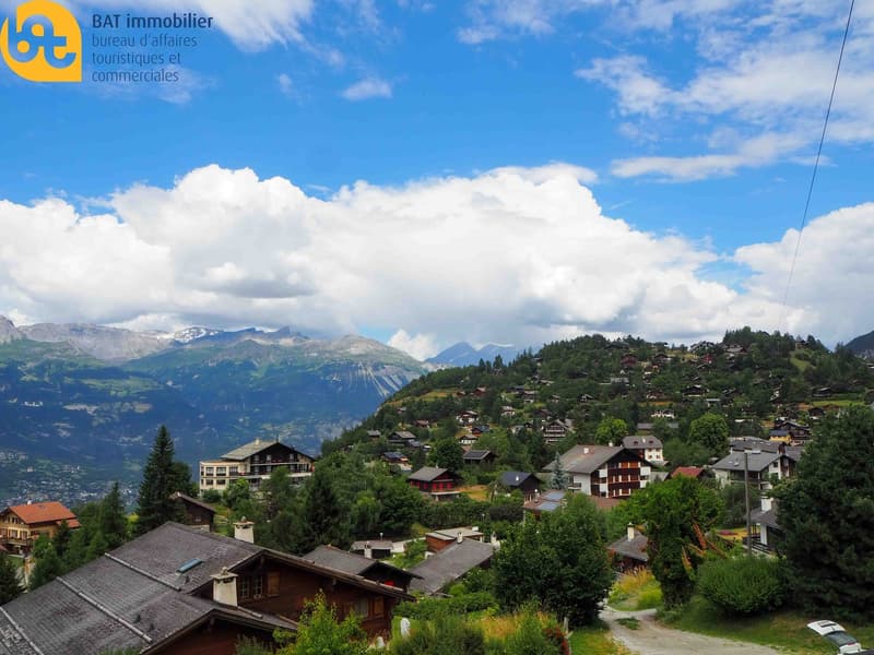 The image shows a picturesque mountain village nestled in a lush, green landscape. The village is surrounded by towering mountains and a blue sky with fluffy white clouds. The houses are traditional Alpine-style buildings with red roofs. There are trees a