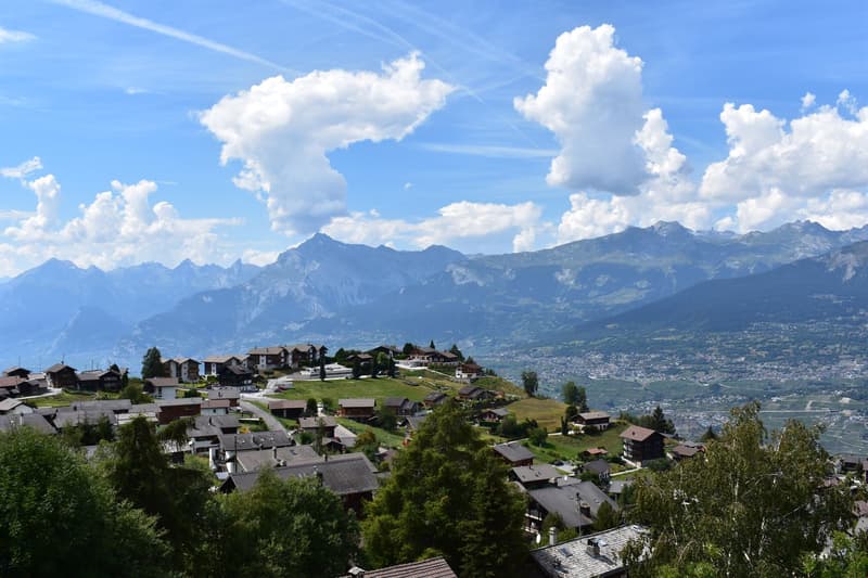 View of a village on a hillside, surrounded by mountains, hills, and trees.