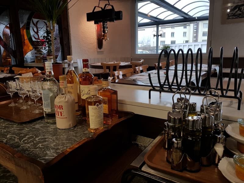 A restaurant counter, wooden tables, chairs, wine glasses, various bottles, a window with a view of buildings, plants, and lamps, a tray with condiments, a marble countertop, and a view of the outside